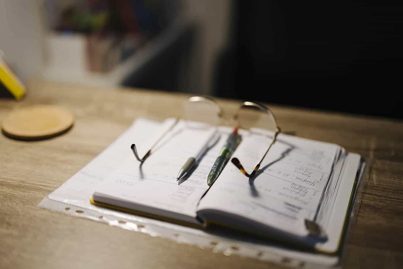 An open notebook with two pens and a pair of glasses on a wooden desk